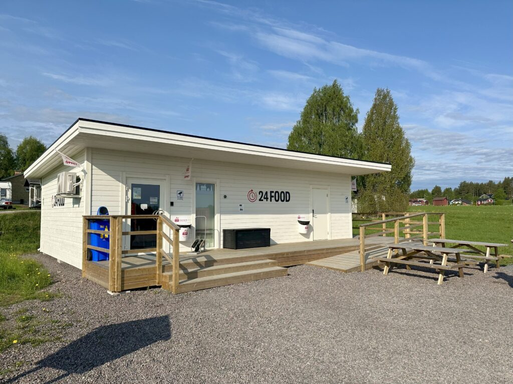 Unbyn En liten, vit, fristående UNEK-byggnad med beteckningen 24FOOD står på en grusplan med träramper, picknickbord, gräsmattor och träd i bakgrunden under en klarblå himmel.