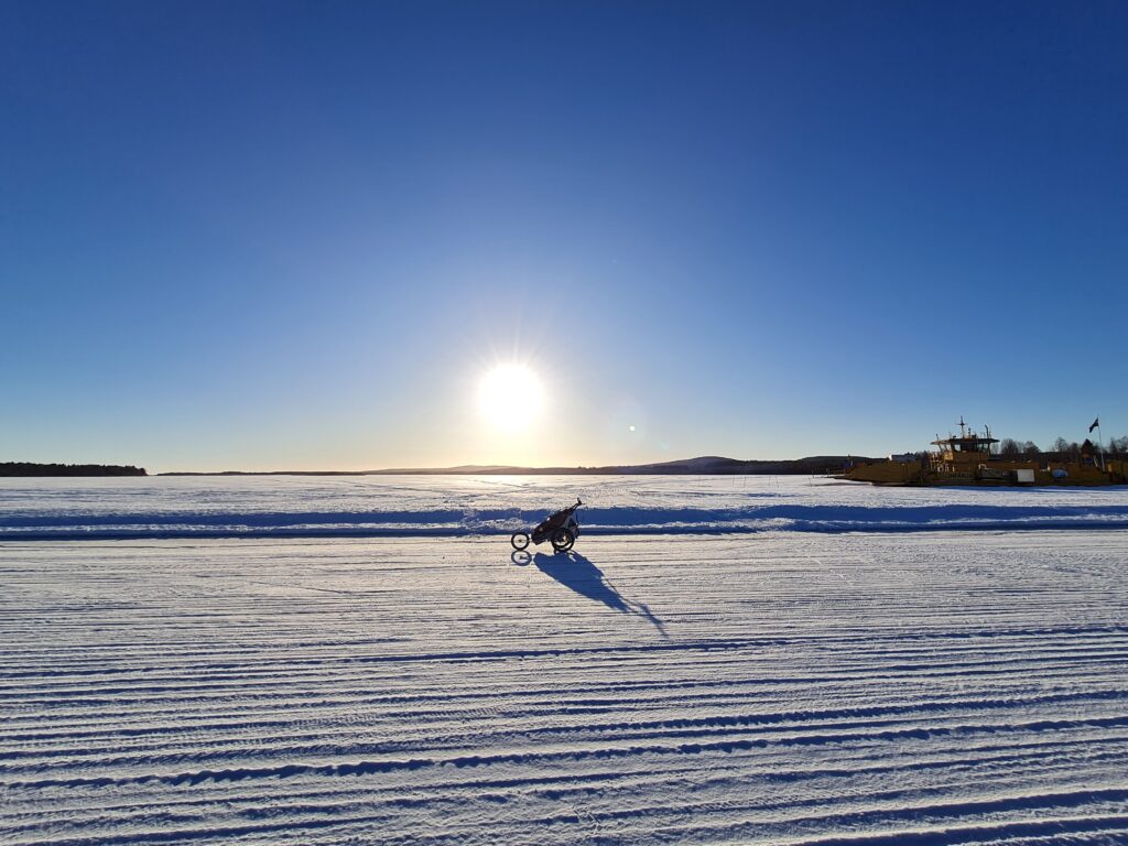 Unbyn En person cyklar på en UNEK-cykel med en liten släde påkopplad genom ett snöigt, fruset landskap under en klarblå himmel, solen skiner lågt vid horisonten.