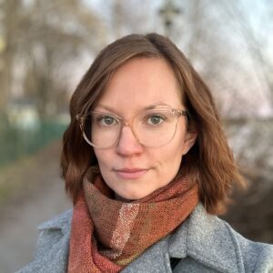 Unbyn En person med kort brunt hår och glasögon bär en grå kappa prydd med en grön och vit rund brosch och en rödbrun halsduk, och står utomhus på en suddig stig med träd i bakgrunden. Den avslappnade hållningen tyder på att personen kanske väntar på någon eller är försjunken i tankar, kanske på väg att ta kontakt – kontakt – med en vän i närheten.